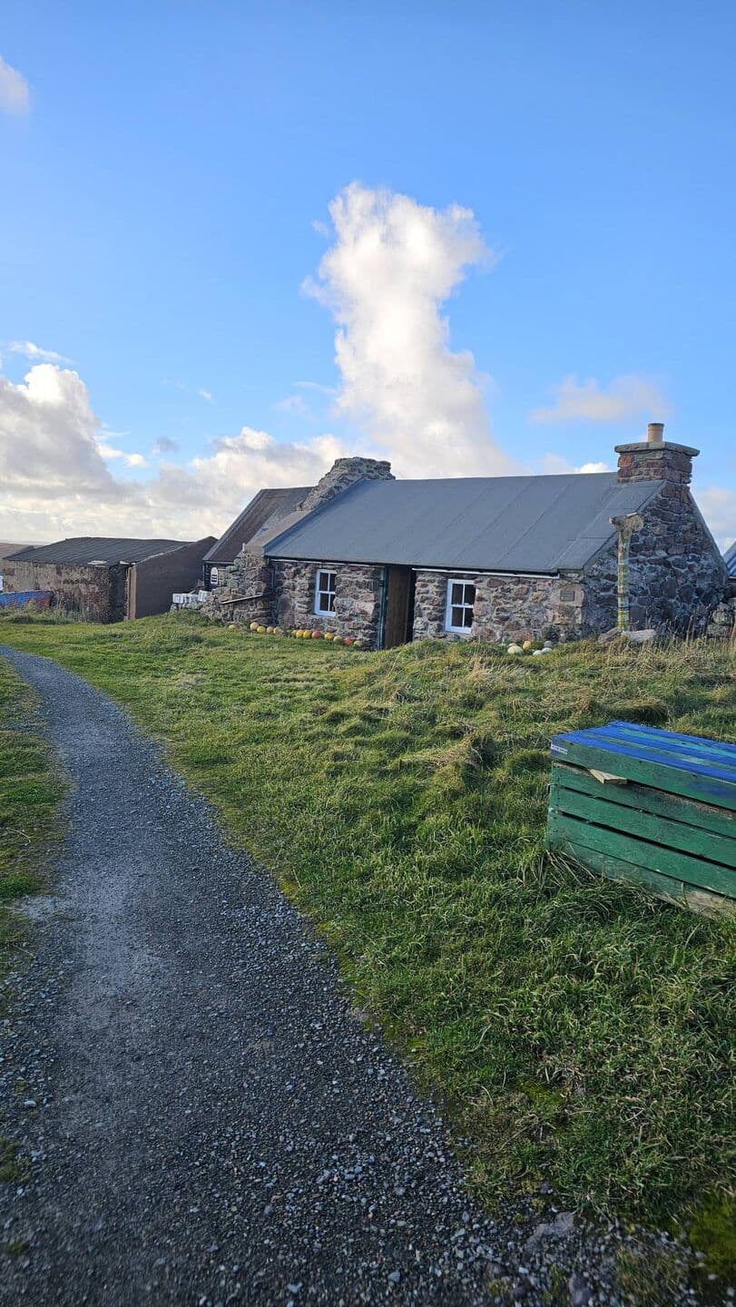 Johnnie Notions Camping Böd from the gravel track: a low stone cottage with white sash windows, a grey roof and fishing floats along the wall