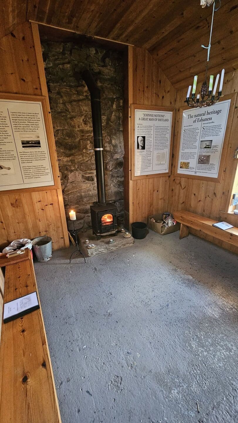 Inside the böd: a wood-burning stove set into a stone wall, with benches and information panels about Johnnie Notions and the natural heritage of Eshaness